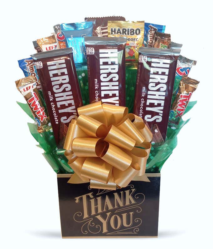 A gift box featuring a variety of candy bars, including Hershey's milk chocolate, Twix, KitKat, Snickers, and Haribo gummy bears, arranged with green wrapping and topped with a large gold bow. The black gift box has a 