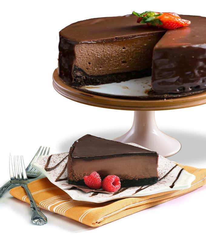 A rich chocolate cheesecake on a pedestal cake stand with a slice cut out and placed on a plate. The cheesecake is topped with glossy chocolate ganache and a strawberry. The slice on the plate is garnished with fresh raspberries and drizzled with chocolat