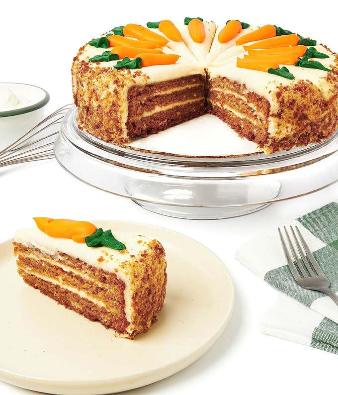 A carrot cake on a glass cake stand with a slice cut out and placed on a plate. The cake is layered with cream cheese frosting and decorated with icing carrots on top. A bowl of extra frosting and a fork are also visible.