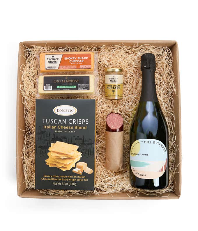 A gift box with Tuscan crisps, salami, cheeses, mustard, and a bottle of Hill & Harbor sparkling wine. Surrounding the box are plates with cheese, salami, mustard, and two glasses of sparkling wine.