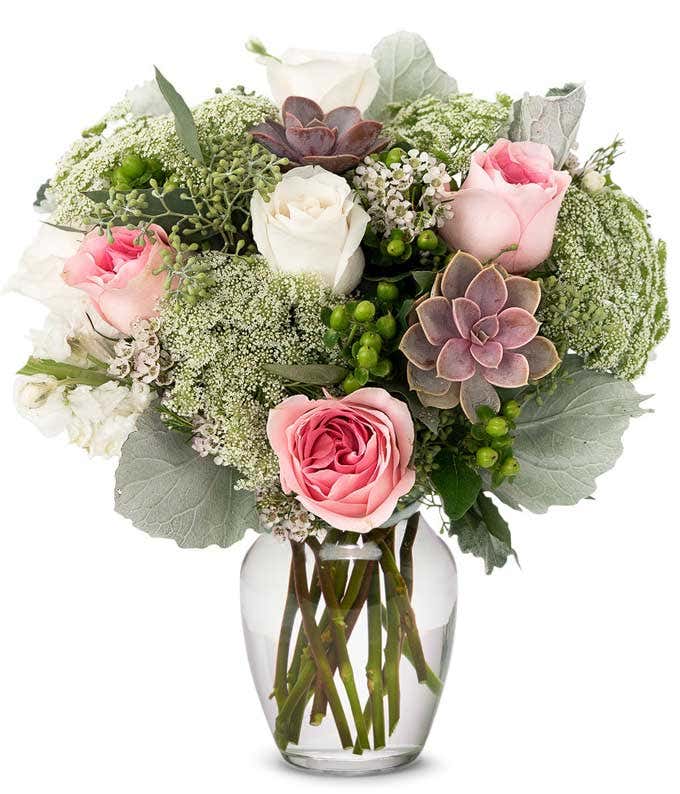 A floral arrangement featuring white roses, pink roses, succulents, white stock, hypericum berries, Queen Anne's lace, white waxflower, and seeded eucalyptus, with an optional glass vase.