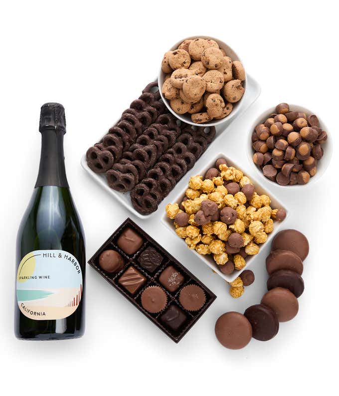 a gift box and a bottle of sparkling wine on a kitchen counter with a glass of wine and some chocolate treats on plates. Gift box includes cookies, chocolates, peanut butter chocolates, chocolate oreos, and chocolate pretzels