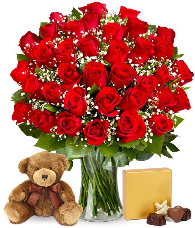 Image of 36 red roses beautifully arranged in a glass cylinder vase, accompanied by a box of chocolates and a cuddly bear plush, the ideal gift for conveying love and warmth.