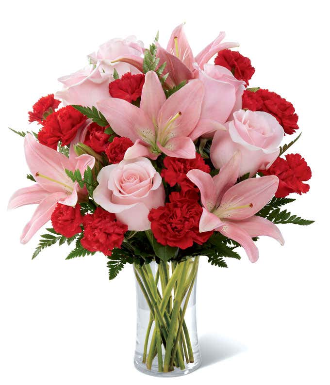 Floral arrangement featuring pink lilies, pink roses, red carnations, and red mini carnations in a cylinder vase.