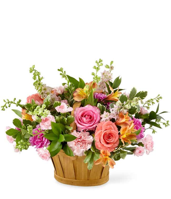 A floral arrangement featuring pink and coral roses (deluxe and premium sizes), pink carnations, pink mini carnations, orange alstroemeria, and pink larkspur, arranged in a wooden basket.