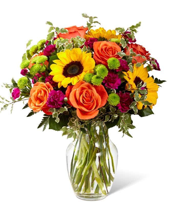 A floral arrangement featuring orange roses, sunflowers, violet mini carnations, and green button poms in a clear vase.