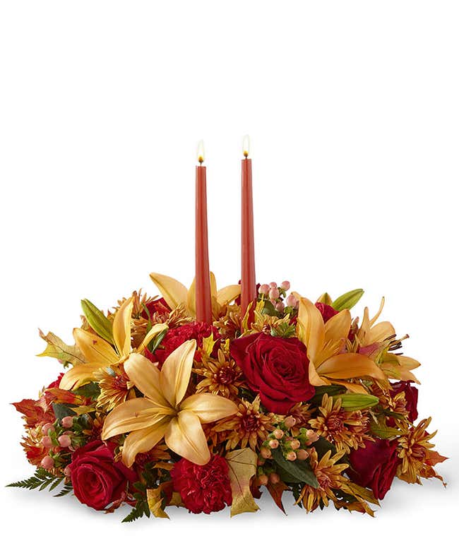 A stunning floral centerpiece featuring red roses, red carnations, orange lilies, peach berries, and bronze cushion poms, complemented by two orange taper candles.