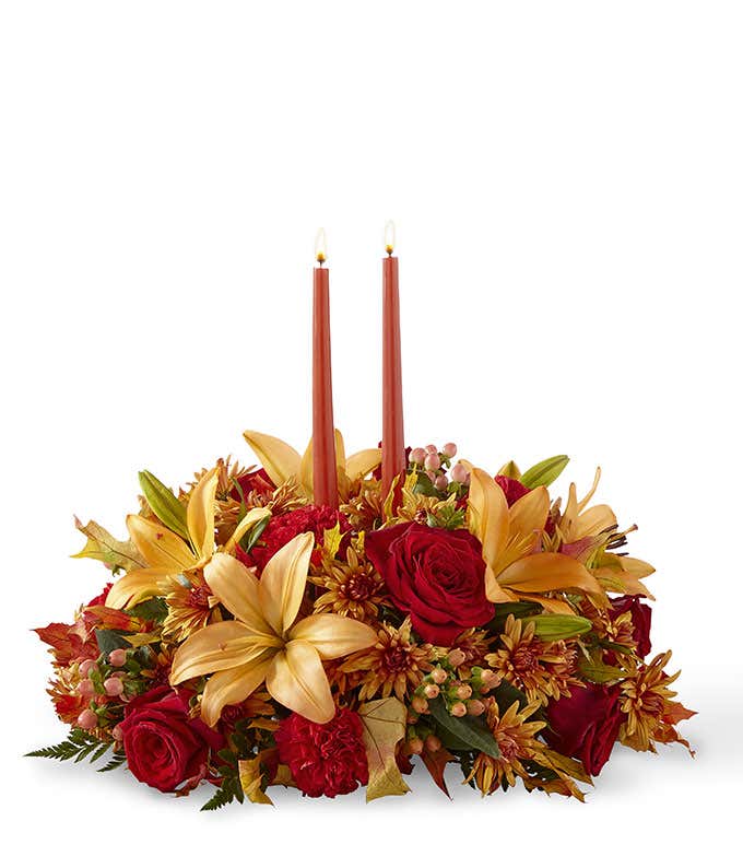 A stunning floral centerpiece featuring red roses, red carnations, orange lilies, peach berries, and bronze cushion poms, complemented by two orange taper candles.