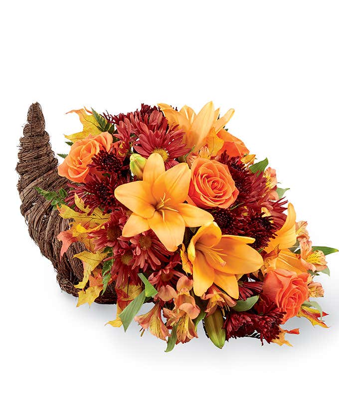 A festive floral arrangement featuring orange lilies, orange roses, orange alstroemeria, and bronze daisies and poms, all beautifully displayed in a cornucopia basket.