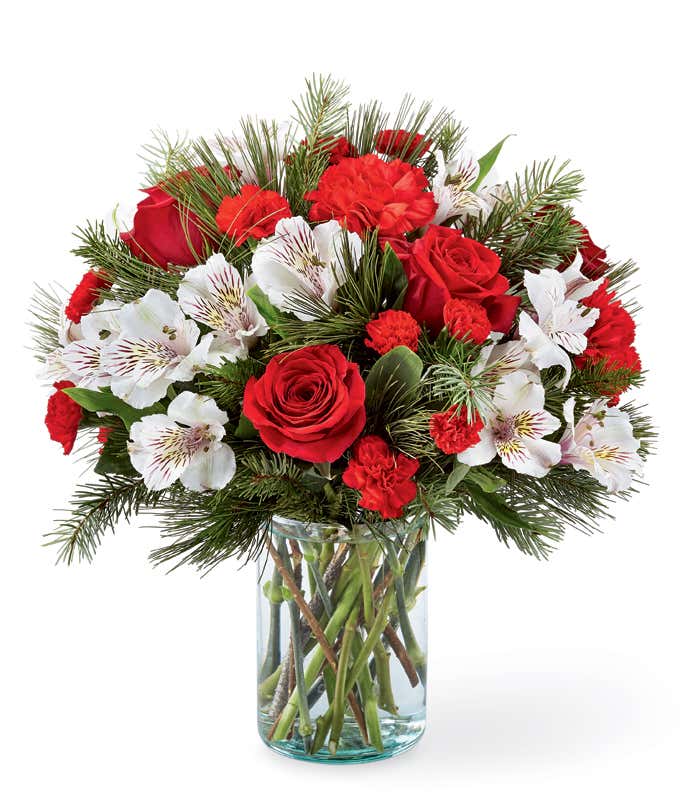 A festive arrangement with red roses, red carnations, red mini carnations, white alstroemeria, and evergreens, beautifully displayed in a glass vase.