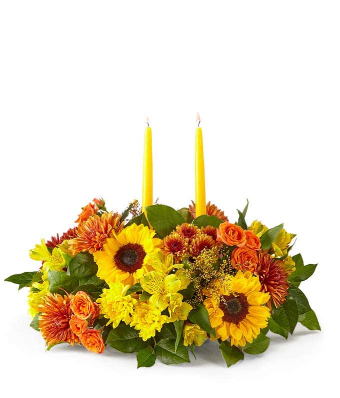 A cheerful floral arrangement featuring sunflowers, butterscotch mums, yellow alstroemeria, butterscotch cushion poms, orange spray roses, yellow cushion poms, red-tinted solidago, and seasonal greens, complemented by yellow taper candles.