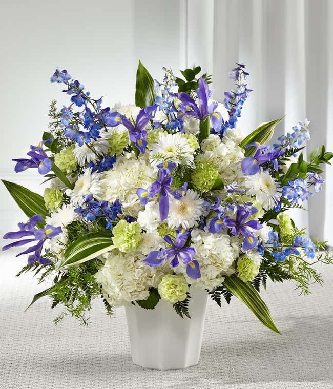 A standing basket of white hydrangeas, blue delphinium, iris, white poms, green carnations, and fresh floral greens in a white plastic urn