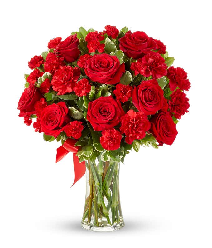 Floral arrangement featuring red roses, red carnations, red mini carnations, and a decorative red ribbon, florist delivered in a clear glass vase.