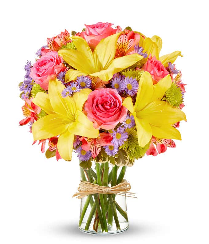 A vibrant arrangement featuring yellow lilies, pink roses, pink alstroemeria, green poms, and purple Monte Casino, beautifully arranged in a glass vase. This colorful mix offers a cheerful and eye-catching display, perfect for brightening any occasion.