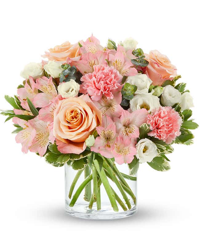 A delightful floral arrangement of peach roses, pink carnations, pink alstroemeria, and white lisianthus, elegantly arranged in a glass vase. This soft and romantic mix of flowers offers a gentle and inviting display.