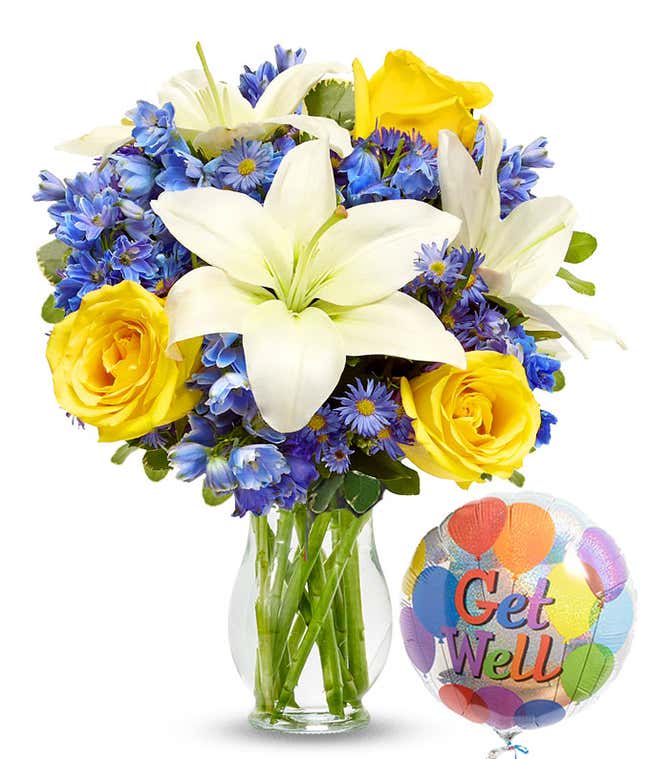 A floral arrangement featuring white Asiatic lilies, yellow roses, blue delphinium, and purple Monte Casino in a keepsake glass vase, accompanied by a 'Get Well' mylar balloon.