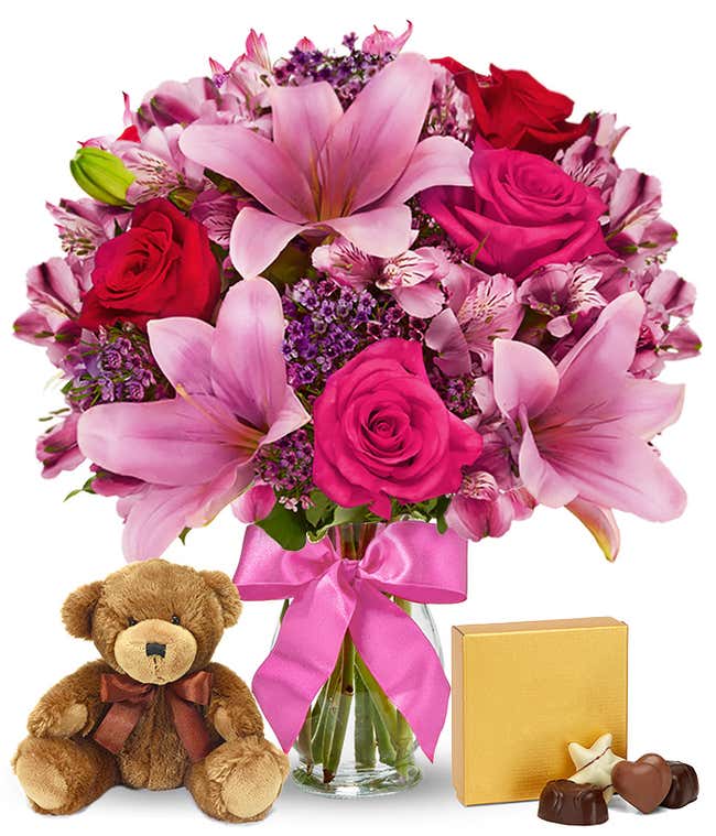 A romantic arrangement featuring red and pink roses, pink lilies, purple waxflowers, and alstroemeria, beautifully arranged in a clear vase with a bow. Includes a keepsake teddy bear and a box of chocolate for an extra touch of love.