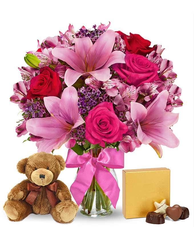 A romantic arrangement featuring red and pink roses, pink lilies, purple waxflowers, and alstroemeria, beautifully arranged in a clear vase with a bow. Includes a keepsake teddy bear and a box of chocolate for an extra touch of love.