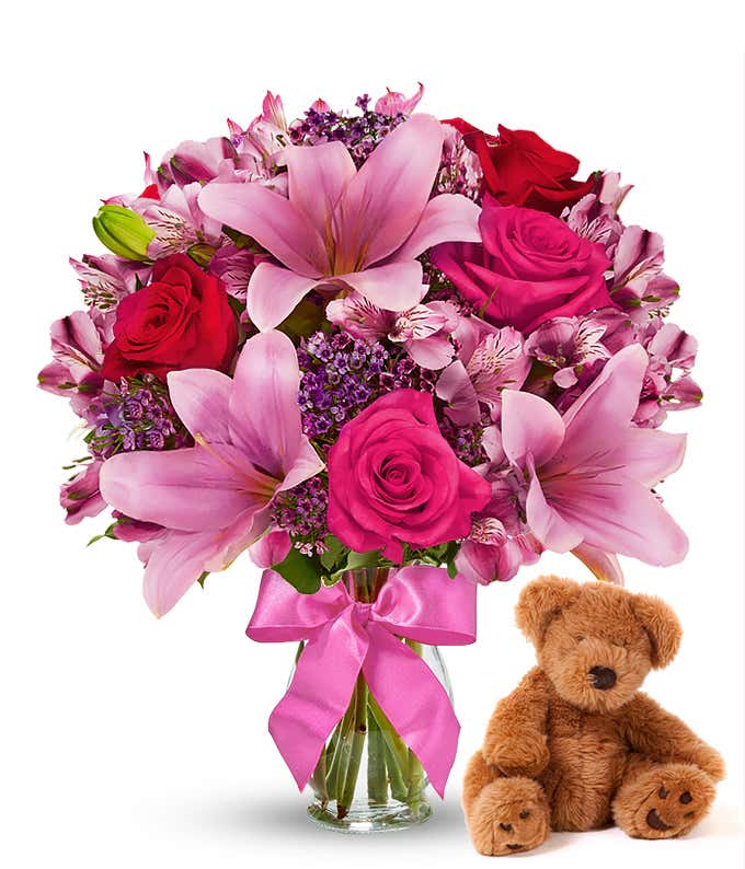 Red and pink roses, pink alstroemeria in a clear vase with bow, accompanied by a keepsake teddy bear.