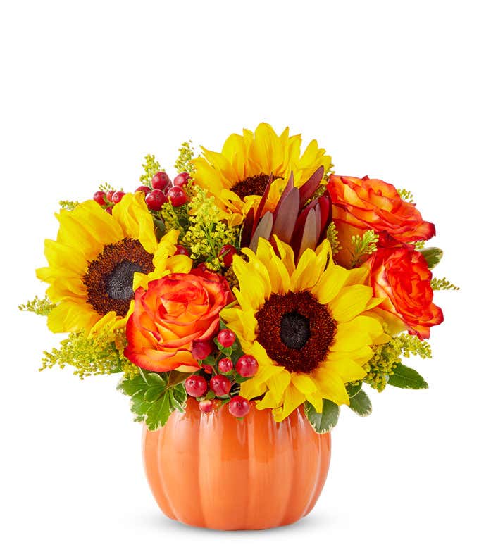  A cheerful floral arrangement featuring sunflowers, orange roses, safari sunset, red berries, and yellow solidago, all beautifully arranged in a pumpkin container.