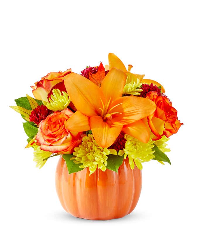  A cheerful floral arrangement featuring orange roses, orange lilies, red Matsumoto asters, and yellow poms, all beautifully arranged in a pumpkin ceramic container.