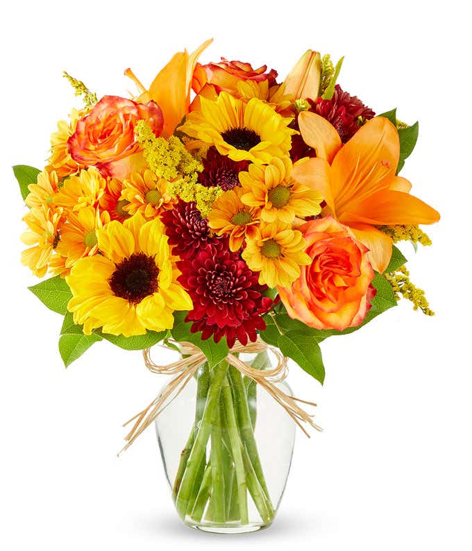 A vibrant fall bouquet in clear glass vase, sunflowers and orange lilies and roses are surrounded by yellow and maroon filler flowers in a clear glass vase.