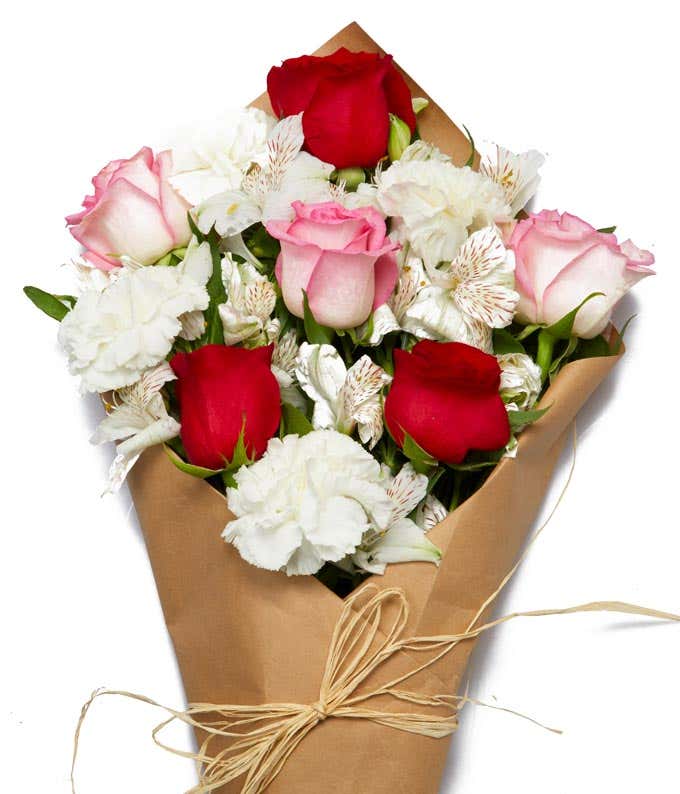 Floral arrangement featuring red roses, pink roses, white carnations, white alstroemerias, fresh greens, wrapped in brown parchment paper with a twine tie.