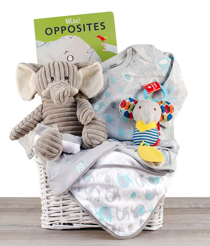 A white basket with a plush elephant toy, a book titled 'Wild Opposites', a cozy blanket, a patterned onesie, and a colorful rattle.