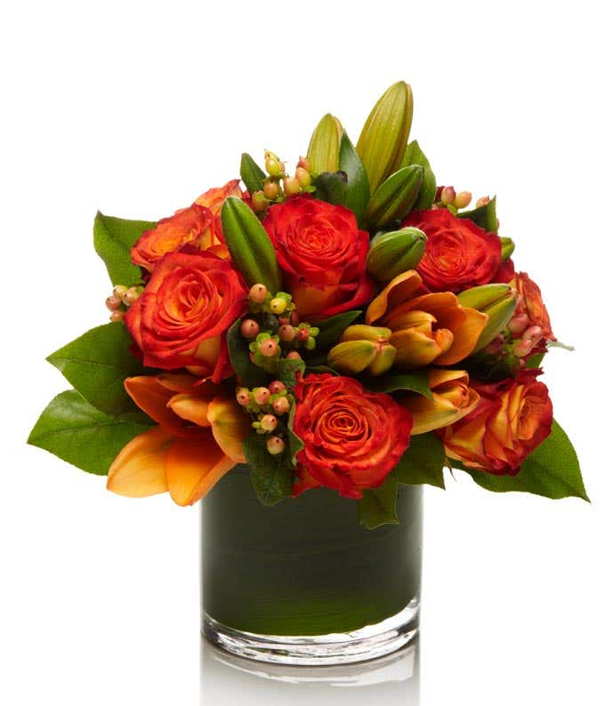 A beautiful floral arrangement featuring orange roses, peach roses, and coral hypericum, all elegantly presented in a round glass vase.