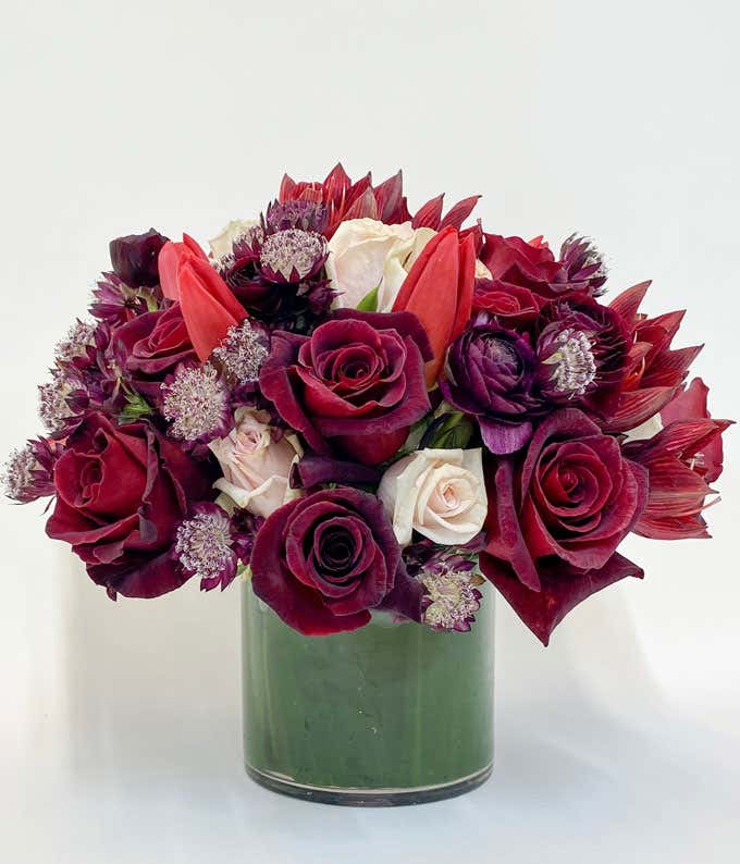 Luxurious bouquet in a clear glass vase featuring a mix of deep red, burgundy, and white flowers, including roses, tulips, and chrysanthemums, arranged with greenery for a rich and elegant floral display.