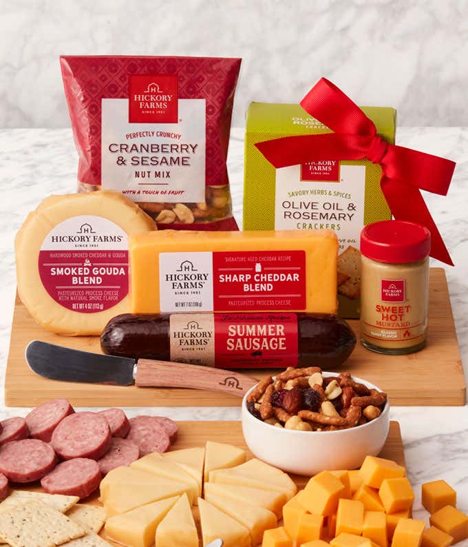 Gourmet gift set with Sharp Cheddar, Smoked Gouda, Olive Oil & Rosemary Crackers, Cranberry & Sesame Nut Mix, Farmhouse Summer Sausage, and Sweet Hot Mustard, displayed on a bamboo cutting board with a cheese spreader.