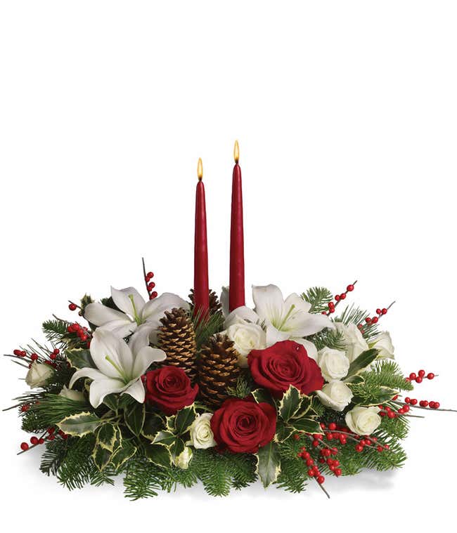 Festive holiday centerpiece featuring red roses, white lilies, white spray roses, red berry accents, evergreen sprigs, and holly, arranged around two tall red taper candles with gold pinecones for a classic seasonal look.