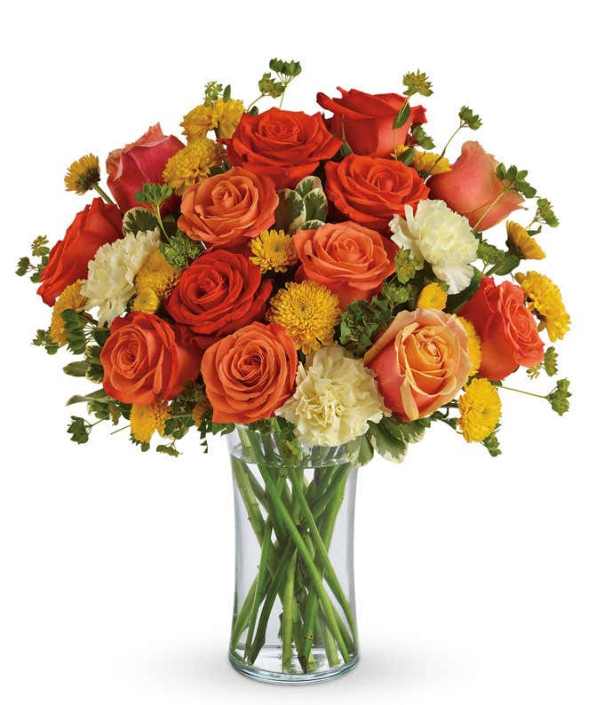 A vibrant floral arrangement featuring orange roses, coral roses, white carnations, and yellow button mums, all beautifully displayed in a glass gathering vase.