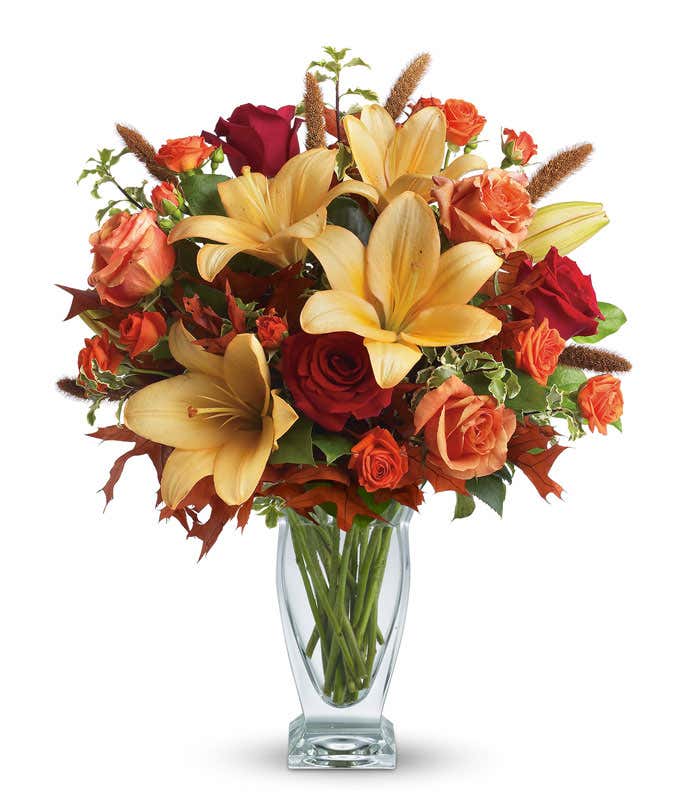 A stunning floral arrangement featuring orange roses, red roses, orange Asiatic lilies, and dried leaves, all elegantly presented in a glass vase.