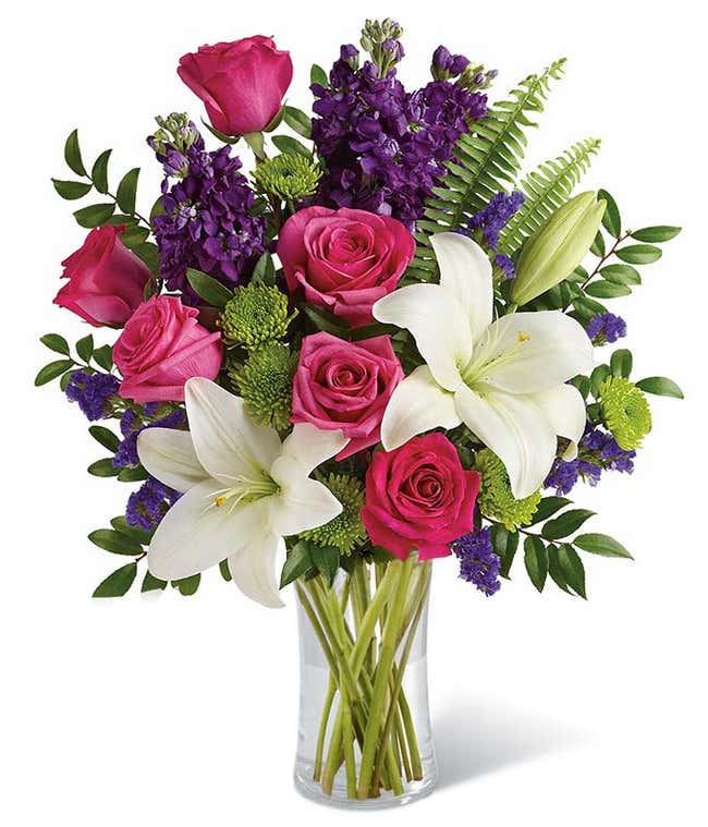 Dark pink roses, white Asiatic lilies, lavender stock, green button spray chrysanthemums, lavender sinuata statice, sword fern, huckleberry in a cylinder vase.