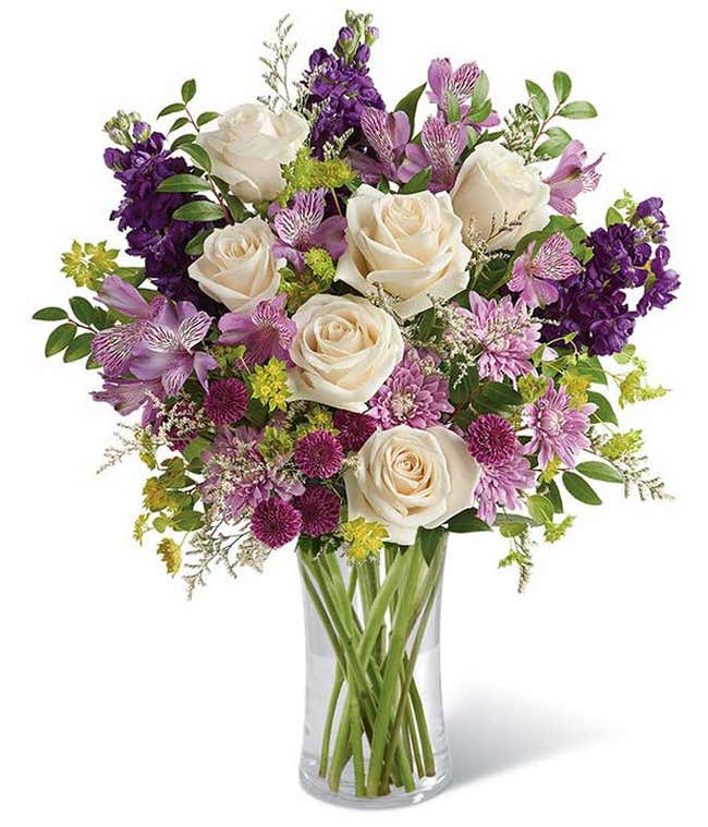 A sophisticated floral arrangement featuring cr&egrave;me roses, lavender alstroemeria, purple stock, purple button chrysanthemums, lavender cushion spray chrysanthemums, white limonium, bupleurum, and huckleberry, all elegantly arranged in a cylinder vase.