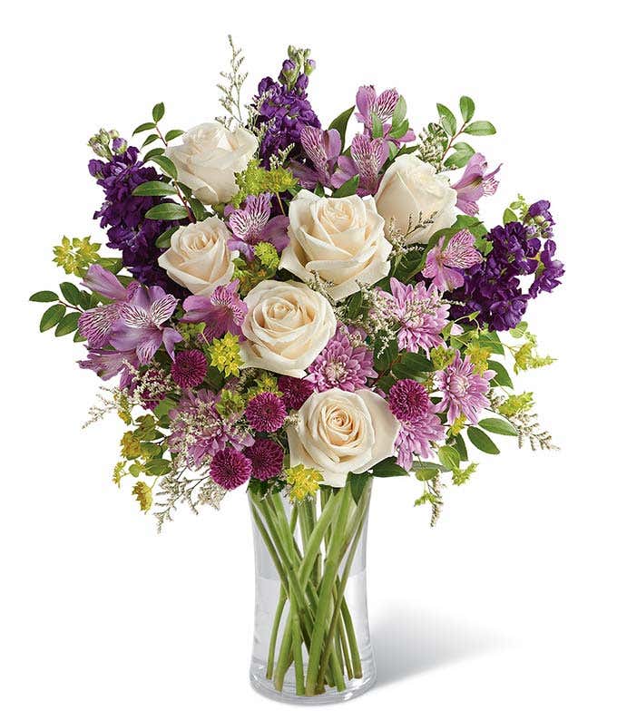 A sophisticated floral arrangement featuring crme roses, lavender alstroemeria, purple stock, purple button chrysanthemums, lavender cushion spray chrysanthemums, white limonium, bupleurum, and huckleberry, all elegantly arranged in a cylinder vase.