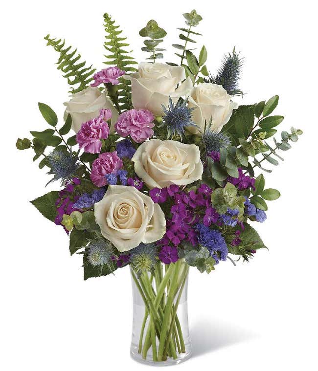 A floral arrangement in a glass cylinder vase featuring cr&egrave;me roses, pink mini carnations, burgundy Sweet William, purple sinuata statice, blue eryngium, huckleberry, spiral eucalyptus, and sword fern.