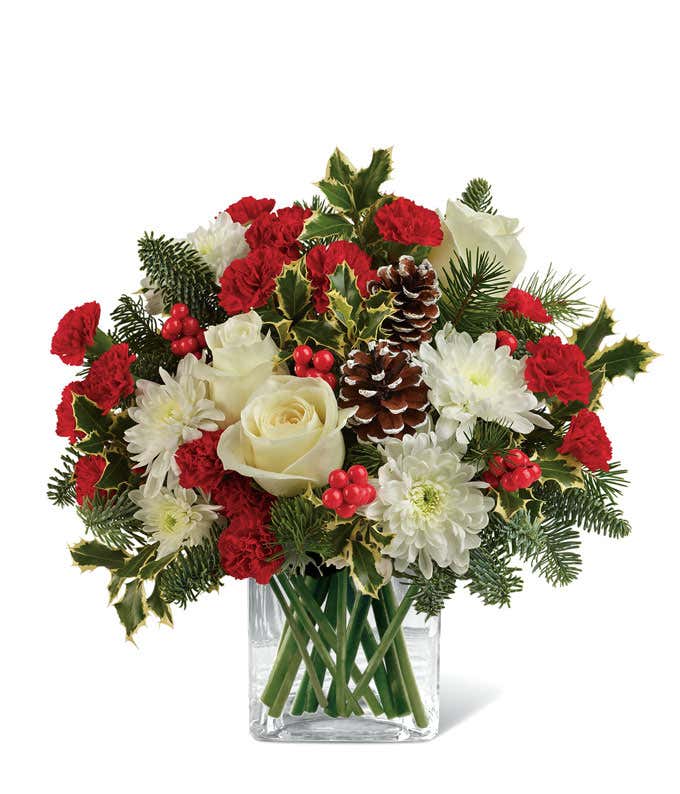 A beautiful floral arrangement with red mini carnations, white roses (deluxe and premium sizes only), white cushion spray chrysanthemums, Douglas fir, variegated holly, noble fir, pinecones, and red berries, displayed in a clear cube vase.