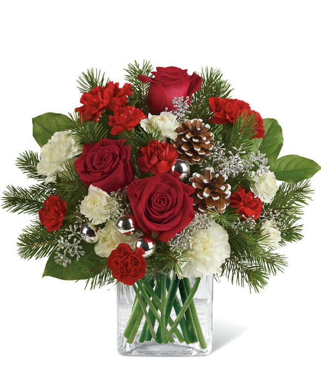 A festive arrangement with red roses (deluxe &amp; premium only), red carnations, white carnations, red mini-carnations, white mini-carnations, seasonal greens, silver ornaments, and pine cones.