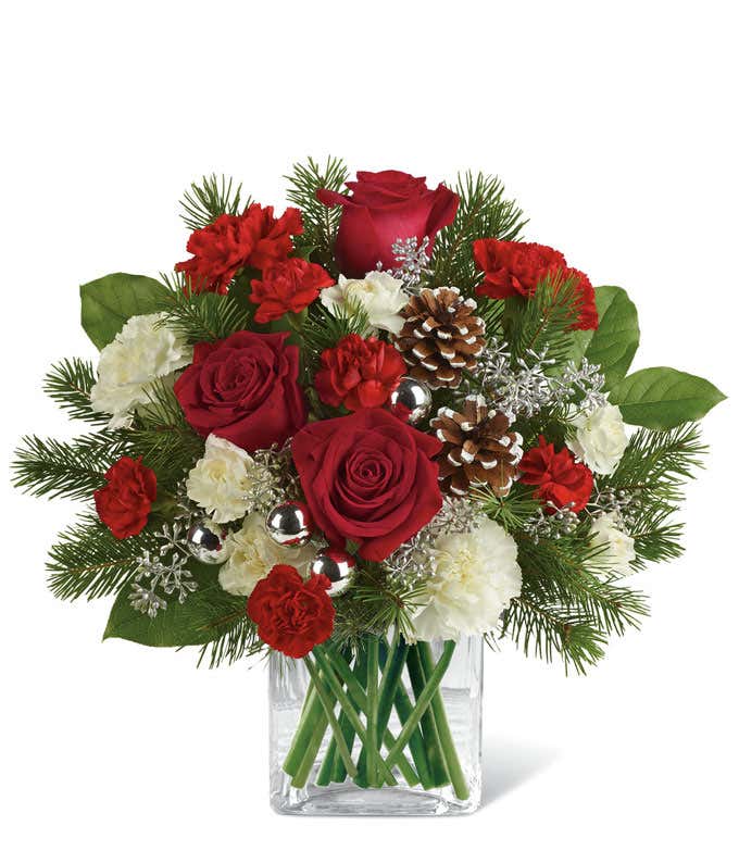 A festive arrangement with red roses (deluxe & premium only), red carnations, white carnations, red mini-carnations, white mini-carnations, seasonal greens, silver ornaments, and pine cones.