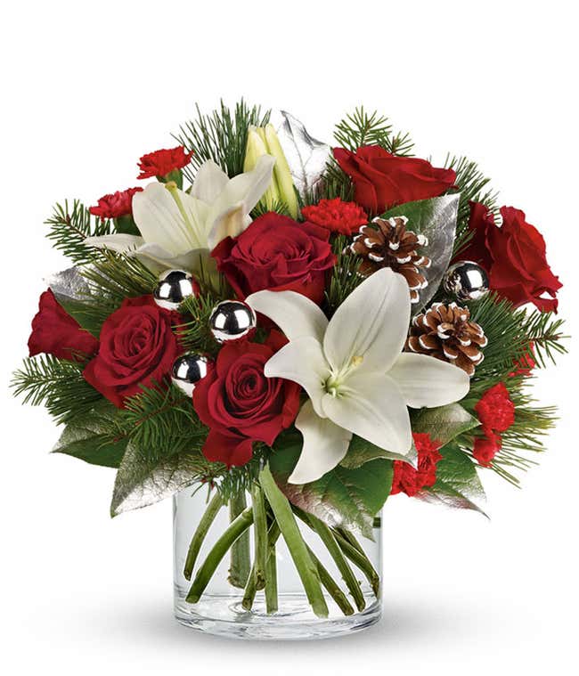 A festive arrangement with red roses, white Asiatic lilies, red mini-carnations, seasonal greens, silver ornaments, and pinecones, beautifully displayed in a clear cylinder vase.