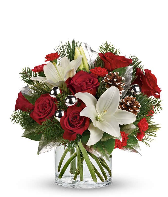 A festive arrangement with red roses, white Asiatic lilies, red mini-carnations, seasonal greens, silver ornaments, and pinecones, beautifully displayed in a clear cylinder vase.