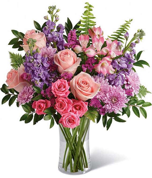 Floral arrangement featuring light pink roses, pink spray roses, pink alstroemeria, lavender stock, lavender mums, raspberry statice, and greenery in a glass vase.