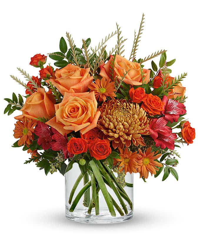 A vibrant floral arrangement featuring orange roses, orange spray roses, red Peruvian lilies, bronze mums, and orange daisies, all elegantly displayed in a clear cylinder vase.