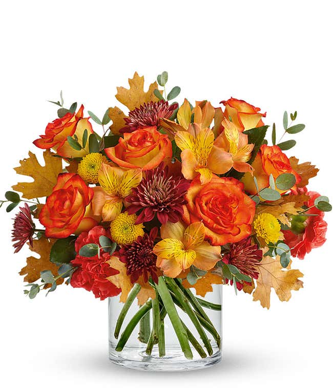 A vibrant fall-themed floral arrangement featuring orange fall roses, orange alstroemeria, yellow button mums, burgundy cushion mums, and orange carnations, all elegantly displayed in a cylinder vase.