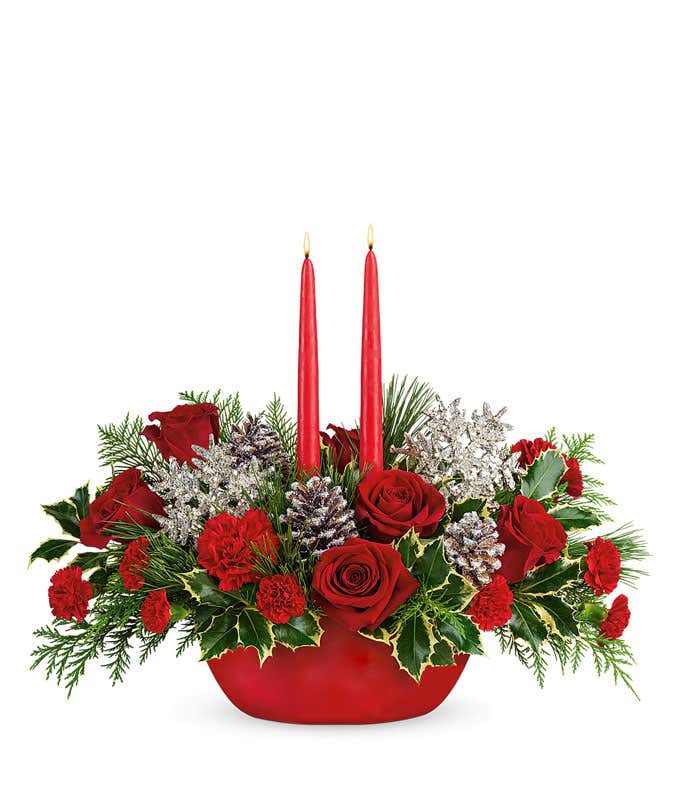 A festive arrangement with red roses, red carnations, red mini carnations, silver snowflakes, snowy pinecones, evergreens, and red candles, displayed in a keepsake container.