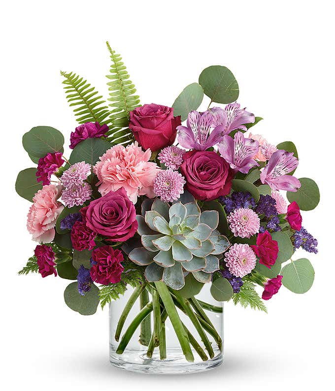 Floral arrangement featuring a succulent plant, pink carnations, purple mini carnations, dark pink roses (in deluxe and premium versions), purple statice, purple alstroemeria, lavender button mums, seasonal greenery, and a personal card message in a glass