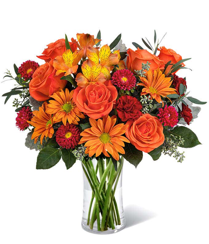 A vibrant floral arrangement featuring orange roses, orange alstroemeria, bronze daisies, red Matsumoto asters, maroon mini carnations, and seasonal greenery, complemented by a personal card message, all beautifully presented in a clear cylinder vase.