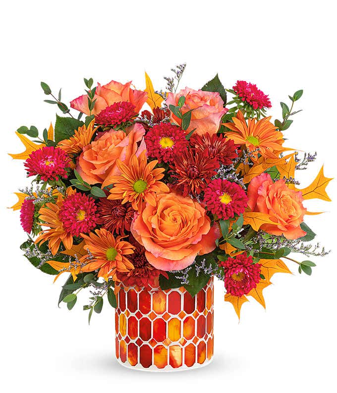 A vibrant floral arrangement featuring orange roses, red Matsumoto asters, bronze mums, and bronze daisies, complemented by greenery and fall leaves. The arrangement includes a personal card message and is elegantly presented in a keepsake vase​